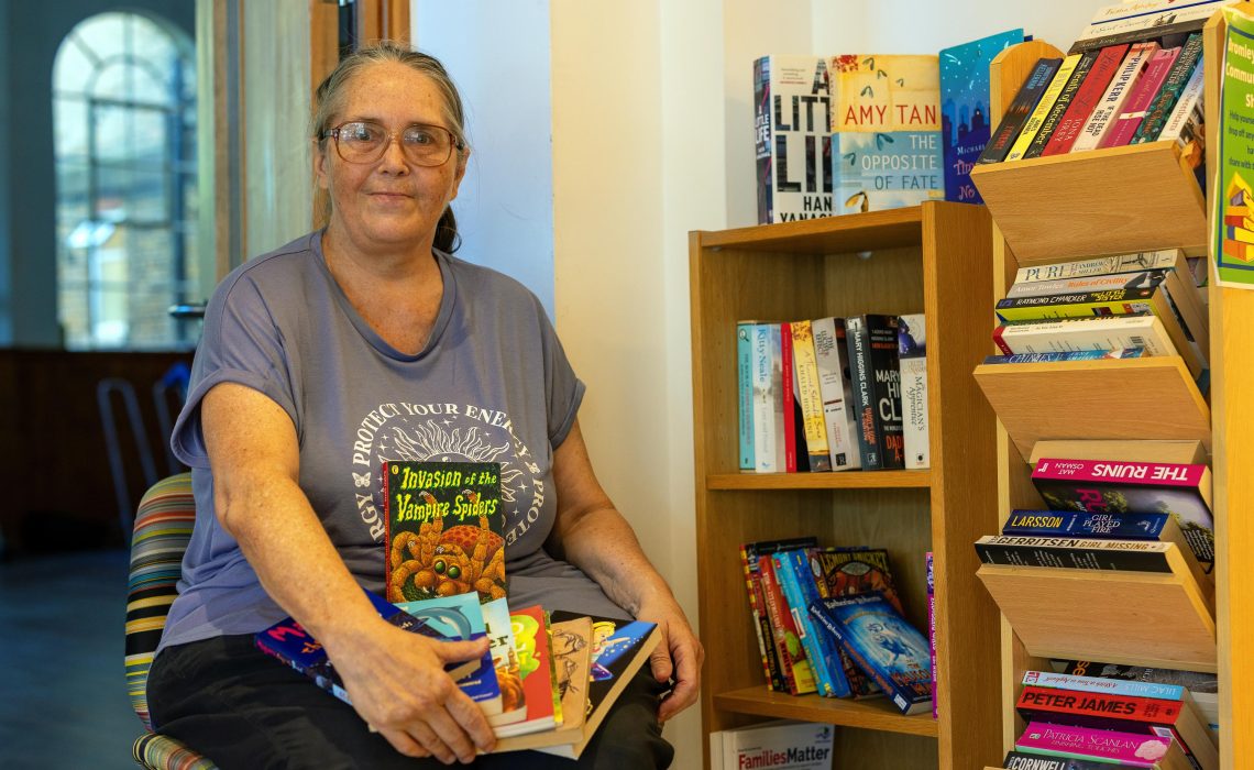 A woman in a grey t-shirt is sat next to a bookcase full of a variety of books